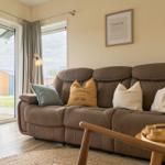 Ein braunes Sofa mit Kissen steht vor einem Fenster. Draußen ist eine Terrasse sichtbar.