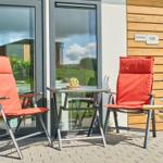 Terrasse mit zwei orangefarbenen Sesseln, Tisch und Grill. Hintergrund: Glasfront und Holzwand.