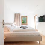 Schlafzimmer mit Bett, Fenstern und Wandfernseher.