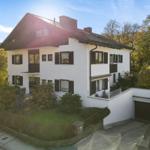 Ein zweistöckiges Haus mit weißen Wänden und dunklen Fensterläden. Umgeben von Grünflächen und Bäumen.