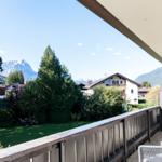 Terrasse mit Blick auf grüne Wiesen und Berge. Geländer und Sitzgelegenheit sichtbar.