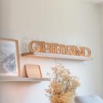 Wandregal mit Holz-Schriftzug und Bilderrahmen. Trockene Blumen im Vordergrund.