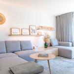 Ein Wohnzimmer mit grauem Sofa, Holztisch und großen Fenstern.