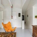 Wohnraum mit braunem Ledersofa, orangefarbenem Stuhl und Holzschrank. Weiße Wände und Deckenbalken.