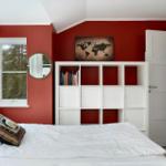 Schlafzimmer mit roten Wänden, weißem Bett und Bücherregal. Fenster mit Blick auf Wald.