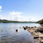 Ruhiger See mit Felsenstrand und bewaldeten Hügeln unter blauem Himmel.