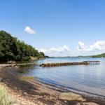 Strand mit Steg und Booten an ruhigem See unter blauem Himmel.