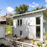 Weiße Holzhütte mit Terrasse, Tisch und Stühlen. Grillhaus und Garten im Hintergrund.