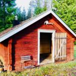 Red wooden house with open door and chair in front. Trees in the background.