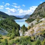 Ein See liegt in einer tiefen Schlucht zwischen bewaldeten Bergen unter blauem Himmel.