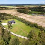 Ein Haus mit Terrasse steht in einer grünen Umgebung mit Feldern und einem See.