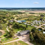 Grüner Campingplatz mit Pool, Wohnwagen und Wald. Blick auf Felder und Windräder.