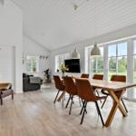 Esszimmer mit Holztisch und braunen Stühlen. Große Fenster zeigen Grünland. Wohnbereich im Hintergrund.