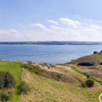 Weitläufiger Blick auf Küstenlandschaft mit Grünflächen und Wasser unter blauem Himmel.