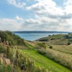 Modernes Haus mit Blick auf ruhige Küstenlandschaft und grüne Hügel unter blauem Himmel.