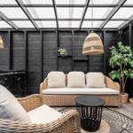 Terrasse mit Rattanmöbeln, Pflanzen und hängenden Lampen unter Glasdach.