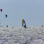 Multiple kitesurfers and windsurfers on the water with colorful sails.