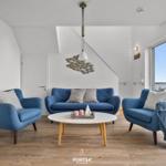Wohnzimmer mit blauer Couch und zwei Sesseln. Blick auf Wasser durch große Fenster.