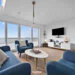 Wohnzimmer mit blauen Sesseln, TV und Meerblick durch große Fenster.
