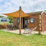Holzhütte mit Terrasse, Sonnensegel und Gartenmöbeln. Hintergrund mit Wohnmobil und Zelt.