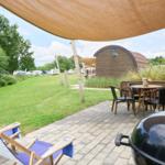 Terrasse mit Sitzgruppe, Grill und Sonnensegel. Grünfläche und Hütte im Hintergrund.