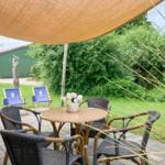 Terrasse mit Tisch, Stühlen, Sonnensegel und Grill. Hintergrund: grüner Garten und Gebäude.