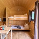 Holzvertäfelte Unterkunft mit Doppelstockbett und Tisch mit Stühlen. Durchgang zur Terrasse.