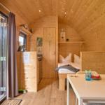 Holzverkleidete Unterkunft mit Schlafplatz, Esstisch und Terrasse mit Blick auf Campingplatz.