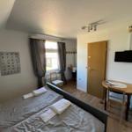 Zimmer mit Doppelbett, Schreibtisch und TV. Fenster mit Vorhängen. Flur mit Tür.