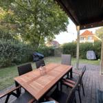 Terrasse mit Tisch und Stühlen, umgeben von Grün und Zäunen.