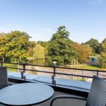 Terrasse mit Tisch und Stühlen, Blick auf einen See und Bäume.
