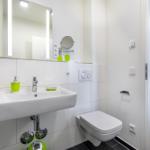Badezimmer mit Waschbecken, Spiegel und Toilette. Grüne Accessoires sichtbar.