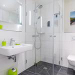 Badezimmer mit Dusche, Waschbecken und WC. Grüne Accessoires und Strandbild.