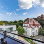 Blick vom Balkon auf Wohnhäuser und Bäume unter blauem Himmel mit weißen Wolken.