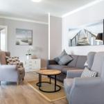 Wohnzimmer mit Sofa, zwei Sesseln und Holztisch. Wandbilder und Küchenbereich sichtbar.