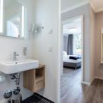 Badezimmer mit Waschbecken und Spiegel. Blick in Schlafzimmer mit Bett und Fenster.