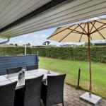 Terrasse mit Tisch, Stühlen und Sonnenschirm. Grünfläche und Hecke im Hintergrund.