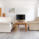 Wohnzimmer mit Sofa, Sessel und Fernseher. Holztisch und große Fenster.