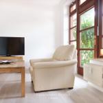 Wohnzimmer mit Sessel, Fernseher und Holztisch. Fenster mit Blick auf Garten.