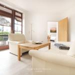 Wohnzimmer mit hellen Möbeln und Holztisch. Durchgang zur Schlafzimmer.