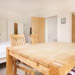 Holztisch mit Stühlen vor Bett und Kleiderschrank. Türen führen zu weiteren Räumen.