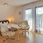 Schlafzimmer mit Bett, Tisch und zwei Stühlen. Große Fenster mit Blick auf Balkon.