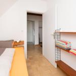 Schlafzimmer mit Bett, Holzregal und Bad im Hintergrund.