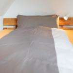 Ein Bett mit Holzkopfteil, grauer Bettwäsche und einer Wandlampe.