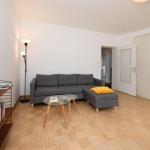 Wohnzimmer mit grauem Sofa, Glas-Tisch und Holzboden. An der Wand steht ein Bücherregal.