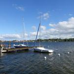 Zwei Segelboote liegen an einem Holzsteg im Wasser. Im Hintergrund sind Bäume und ein klarer Himmel.