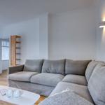 Wohnzimmer mit grauem Sofa, weißen Wänden und Holzboden. Es gibt eine Couchtisch und eine Wandlampe.