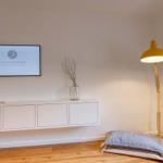 Wohnzimmer mit Wand-TV, weißen Schränken und gelber Stehlampe.
