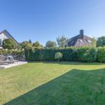 Hinterhof mit Rasenfläche, Holzterrasse und Liegen. Umzäunt mit Hecke. Himmel klar blau.