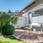 Terrasse mit Sitzgelegenheit, Pflanzen und Garten. Hauswand mit Fenster und Markise.